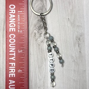May include: A silver keyring with a chain and beaded accents. The keychain features gray and clear beads, with the letters "TTPD" spelled out on white letter beads. A red ruler with the text "ORANGE COUNTY FIRE AUTHO" is on the left.