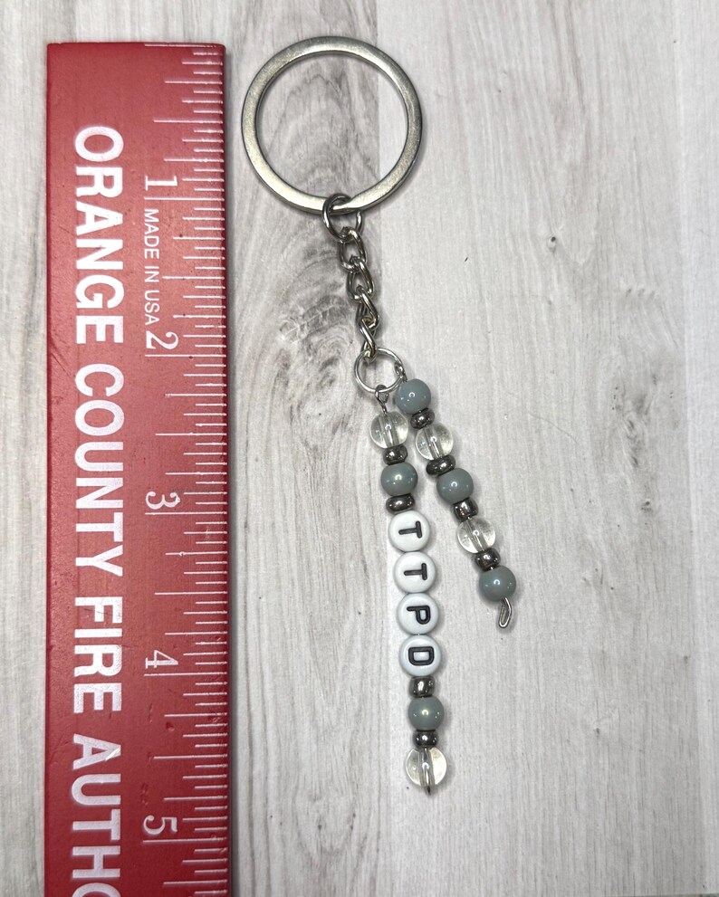 May include: A silver keyring with a chain and beaded accents. The keychain features gray and clear beads, with the letters "TTPD" spelled out on white letter beads. A red ruler with the text "ORANGE COUNTY FIRE AUTHO" is on the left.