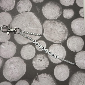 Op de afbeelding: Een kralen sleutelhanger met het woord "FOLKLORE" gespeld in witte letterkralen. De sleutelhanger heeft een zilveren sluiting en is versierd met kleine, zilveren en parelachtige kralen. De achtergrond heeft een patroon van grijze en witte houtschijven.