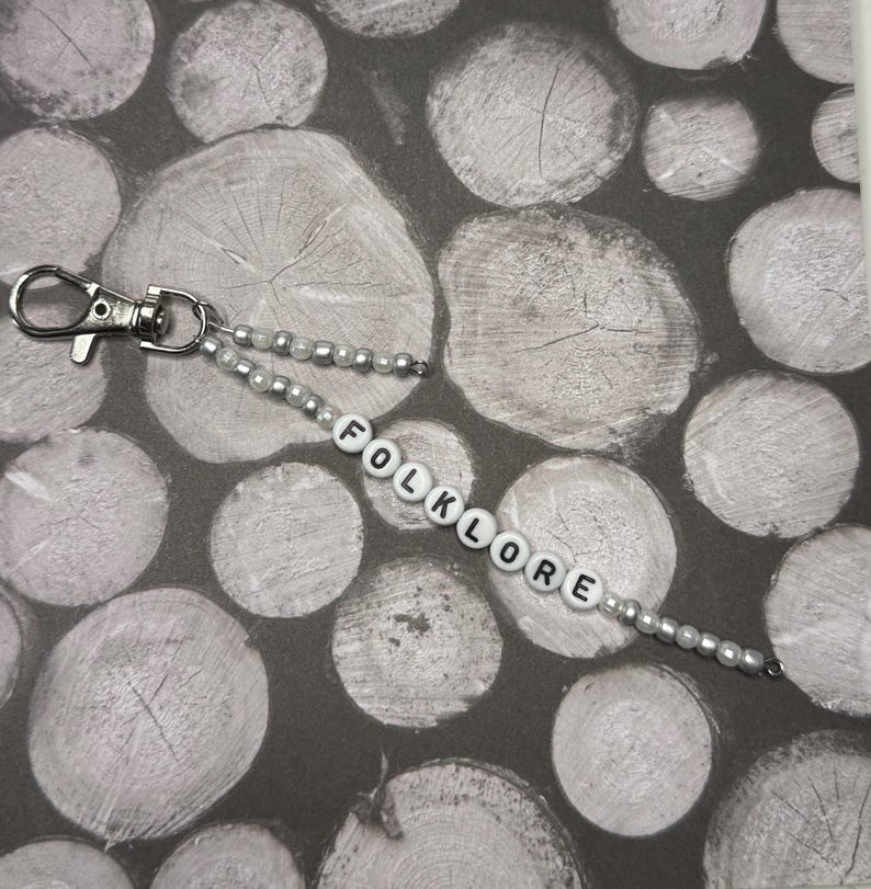 Op de afbeelding: Een kralen sleutelhanger met het woord "FOLKLORE" gespeld in witte letterkralen. De sleutelhanger heeft een zilveren sluiting en is versierd met kleine, zilveren en parelachtige kralen. De achtergrond heeft een patroon van grijze en witte houtschijven.
