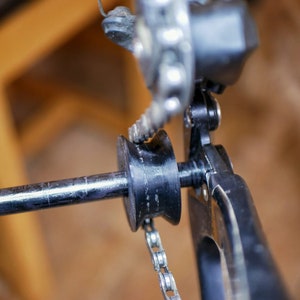 Puede incluir: Primer plano de un desviador de bicicleta con una cadena y una rueda guía de plástico negro. El desviador está unido a un cuadro de metal plateado.