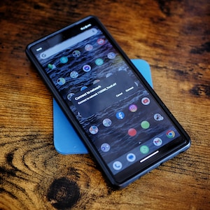 Op de afbeelding: Een zwarte smartphone met een blauwe achtergrond. Het telefoonscherm toont een melding met de tekst "Verbind met netwerk 112300E7300F7004d7". De telefoon heeft verschillende app-pictogrammen op het startscherm.