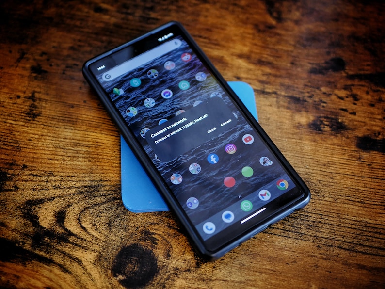 Op de afbeelding: Een zwarte smartphone met een blauwe achtergrond. Het telefoonscherm toont een melding met de tekst "Verbind met netwerk 112300E7300F7004d7". De telefoon heeft verschillende app-pictogrammen op het startscherm.