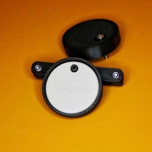 K&ouml;nnte beinhalten: Ein wei&szlig;es rundes Chip-Tracking-Ger&auml;t mit dem Wort "chipolo" darauf gedruckt. Das Ger&auml;t ist an einem schwarzen Kunststoffhalter mit zwei kleinen schwarzen Kn&ouml;pfen befestigt.