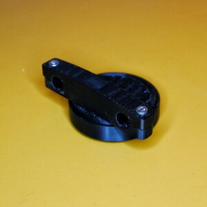 K&ouml;nnte beinhalten: Ein schwarzes, 3D-gedrucktes Objekt aus Kunststoff mit einer runden Basis und einer flachen, rechteckigen Oberseite. Das Objekt hat zwei L&ouml;cher oben und drei silberne Schrauben.