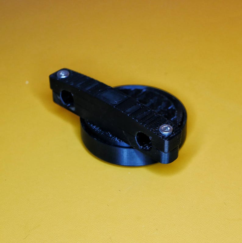 K&ouml;nnte beinhalten: Ein schwarzes, 3D-gedrucktes Objekt aus Kunststoff mit einer runden Basis und einer flachen, rechteckigen Oberseite. Das Objekt hat zwei L&ouml;cher oben und drei silberne Schrauben.