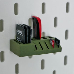 May include: A green storage organizer for SD cards, microSD cards, and a USB drive. The organizer is mounted on a white pegboard. The organizer holds multiple black SD cards with the Sandisk logo and a red and black USB drive.