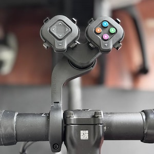 Op de afbeelding: Zwarte Garmin fietsstuurafstandsbediening met twee bedieningseenheden. Eén eenheid heeft pijlen, de andere gekleurde knoppen. De afstandsbediening is gemonteerd op een zwart stuur met een Shimano stuurpen.