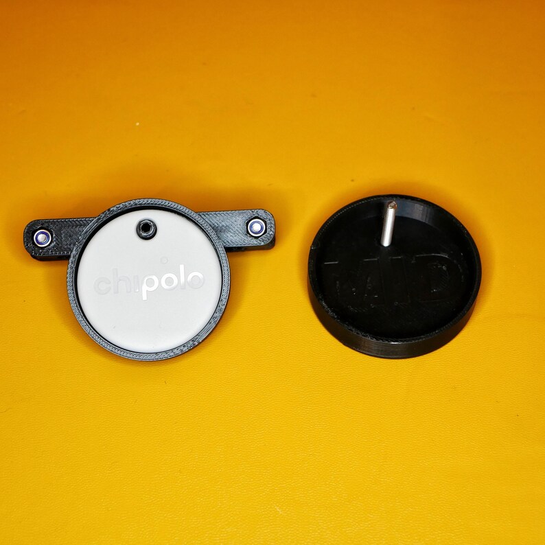 K&ouml;nnte beinhalten: Ein runder wei&szlig;er Sensor mit dem Wort "chipolo" darauf, befestigt an einer schwarzen Kunststoffbasis mit zwei kleinen wei&szlig;en Kn&ouml;pfen. Der Sensor befindet sich neben einer schwarzen Kunststoffbasis mit einem kleinen Metallstift, der aus der Mitte herausragt.