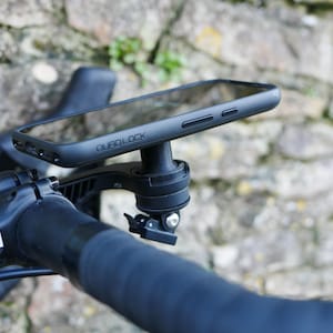 Op de afbeelding: Zwarte Quad Lock telefoonhouder bevestigd aan een fietsstuur. De telefoonhoes is zwart en heeft het Quad Lock logo. Het stuur is zwart met zwart stuurlint. De achtergrond is een stenen muur.