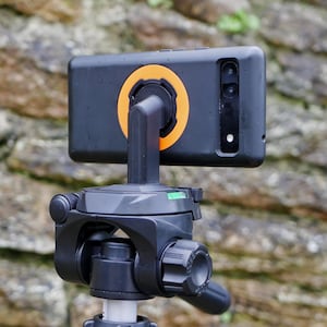 Puede incluir: Un teléfono inteligente negro con un soporte de teléfono negro y naranja conectado a un trípode negro.