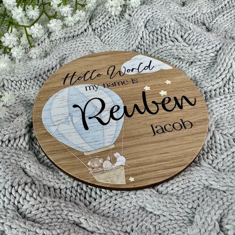 May include: A round wooden sign featuring a blue and white hot air balloon, two bears, and stars. The sign reads "Hello World my name is Reuben Jacob".
