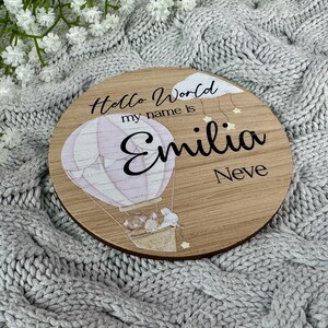 May include: A round wooden sign with a hot air balloon design. The sign says "Hello World my name is Emilia Neve".