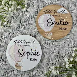 May include: Two wooden round signs with a hot air balloon design. The top sign says "Hello World my name is Emilia Neve". The bottom sign says "Hello World my name is Sophie Rose".