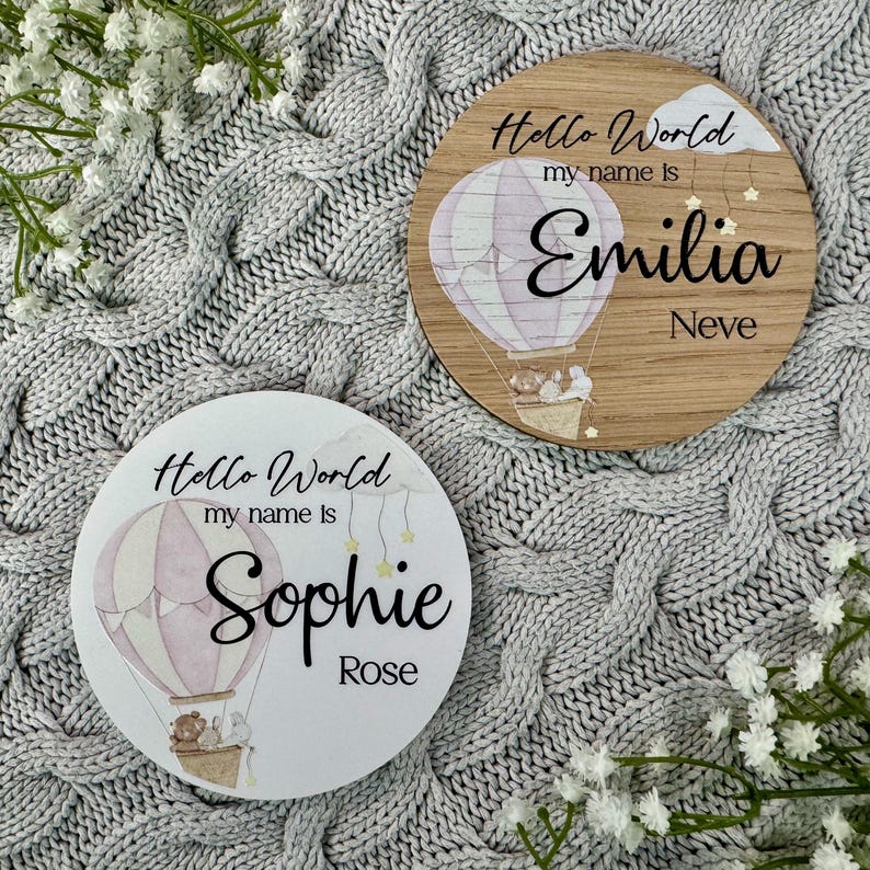May include: Two wooden round signs with a hot air balloon design. The top sign says "Hello World my name is Emilia Neve". The bottom sign says "Hello World my name is Sophie Rose".