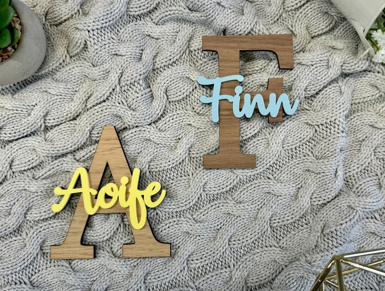 May include: Wooden letter cutouts, "A" with yellow "Aoife" and "F" with light blue "Finn" layered on top. The letters are on a gray cable knit blanket. Decorative items for home decor.