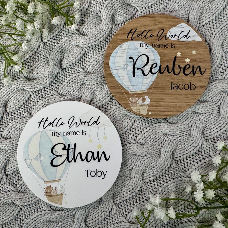 May include: Two round wooden signs with a hot air balloon design. The signs say "Hello World, my name is" and include a name and a second name. The first sign says "Reuben Jacob" and the second sign says "Ethan Toby".
