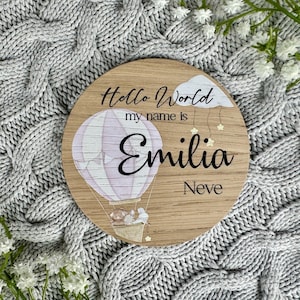 May include: A wooden round sign with a hot air balloon design. The sign says "Hello World my name is Emilia Neve".
