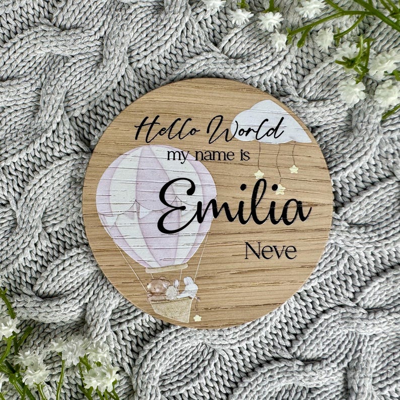May include: A wooden round sign with a hot air balloon design. The sign says "Hello World my name is Emilia Neve".