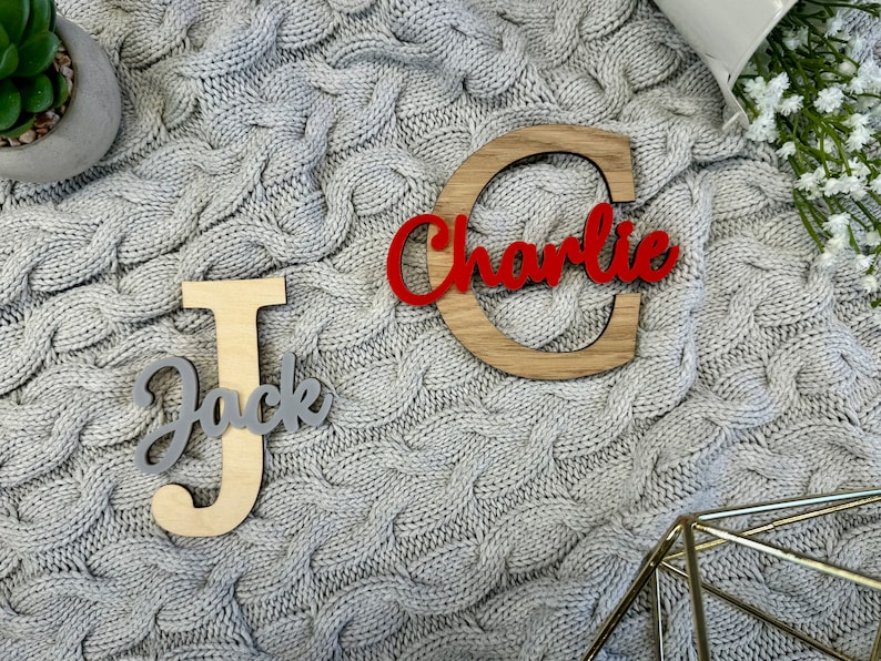 May include: Wooden name cutouts, featuring the names "Jack" and "Charlie". The name "Jack" is in a gray script font on a light wood letter "J". The name "Charlie" is in a red script font on a wooden letter "C".