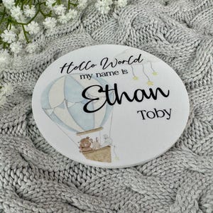 May include: White round wooden sign with a blue and white hot air balloon, two animals, and the text "Hello World my name is Ethan Toby".