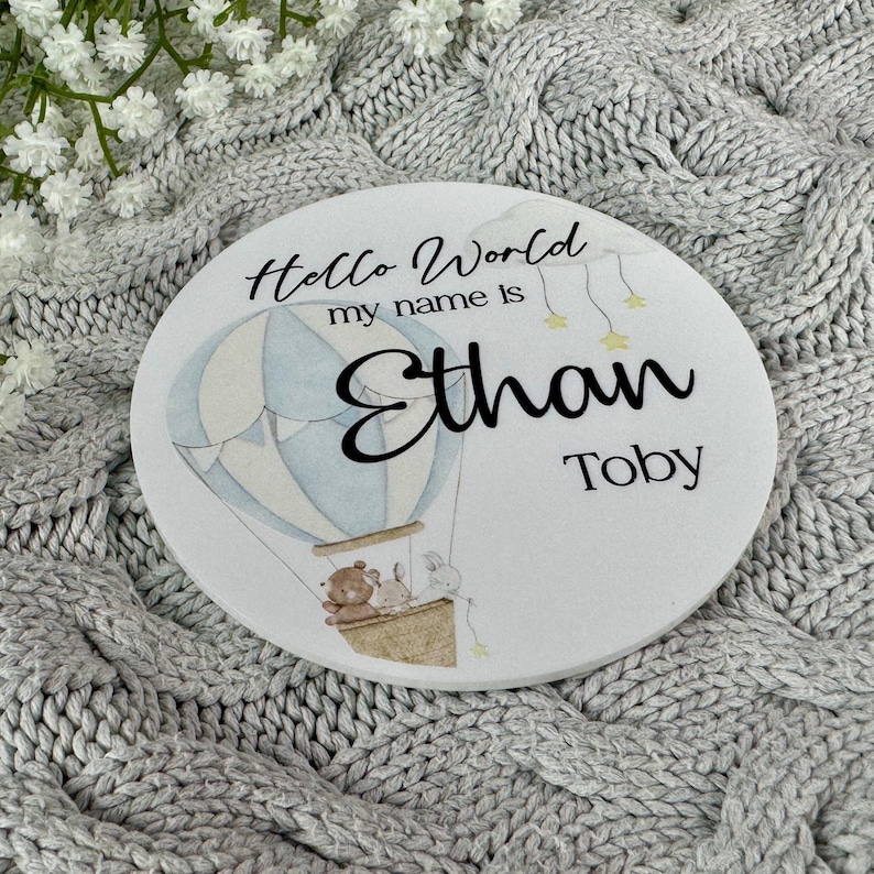 May include: White round wooden sign with a blue and white hot air balloon, two animals, and the text "Hello World my name is Ethan Toby".