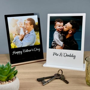 May include: Two black and white photo frames with a wooden base. The frames are in a Polaroid style. The left frame has a photo of a father and son with the text "Happy Father's Day". The right frame has a photo of a father and son with the text "Me & Daddy".