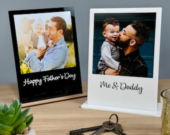 Puede incluir: Dos marcos de fotos en blanco y negro con base de madera. Los marcos son de estilo Polaroid. El marco de la izquierda tiene una foto de un padre y un hijo con el texto "Happy Father's Day". El marco de la derecha tiene una foto de un padre y un hijo con el texto "Me & Daddy".