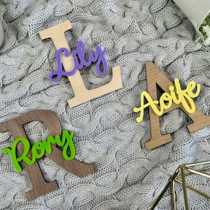 May include: Wooden letter cutouts spelling out the names "Lily", "Aoife", and "Rory". The letters are made of wood in various shades of brown and natural wood, with colorful acrylic name overlays in purple, yellow, and green.