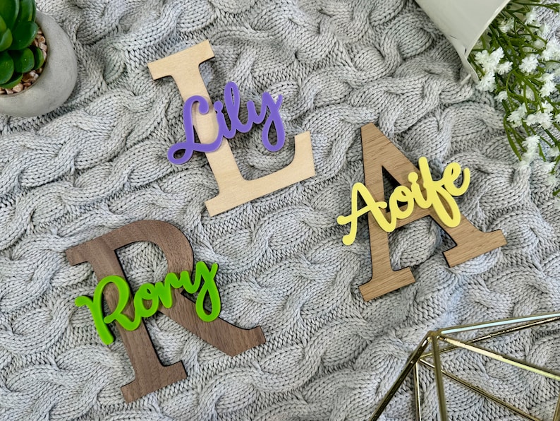 May include: Wooden letter cutouts spelling out the names "Lily", "Aoife", and "Rory". The letters are made of wood in various shades of brown and natural wood, with colorful acrylic name overlays in purple, yellow, and green.