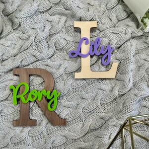 May include: Wooden letter "R" in dark brown with the name "Rory" in green script, and a wooden letter "L" with the name "Lily" in purple script. The letters are on a gray cable-knit blanket.