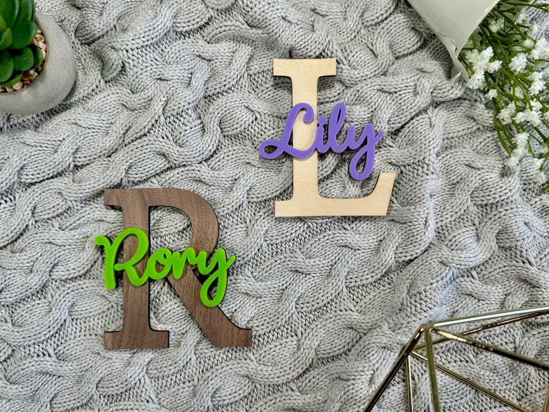 May include: Wooden letter "R" in dark brown with the name "Rory" in green script, and a wooden letter "L" with the name "Lily" in purple script. The letters are on a gray cable-knit blanket.