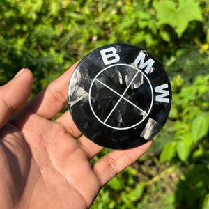 May include: A black and silver BMW emblem with the BMW logo in white. The emblem is round and has a carbon fiber texture. The emblem is being held in a hand against a background of green foliage.