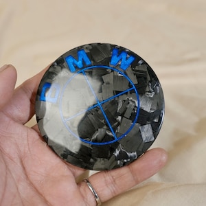 May include: A round, carbon fiber emblem with the BMW logo in blue. The emblem has a textured, dark gray and black pattern. The BMW logo features the letters "B", "M", and "W" in a bold, sans-serif font, with a blue circle and lines.