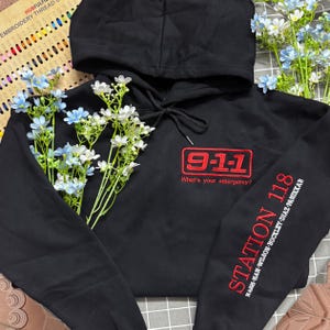 May include: A black hooded sweatshirt featuring red embroidery on the chest that reads 9-1-1 What's your emergency? and red text on the sleeve that reads STATION 118 followed by the names NASH, HAN, WILSON, BUCKLEY, DIAZ, and PANIKKAR.