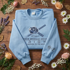 Könnte beinhalten: Hellblaues Sweatshirt mit Rundhalsausschnitt und einem Grafikdesign eines Weltraummoduls mit dem Text "ARTEMIS II", "SPACE MODULE ORION" und "MISSION TO THE MOON". Das Sweatshirt hat ein Design auf dem Ärmel und ist von Blumen umgeben.
