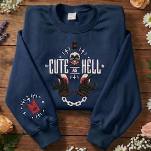 Può includere: Una felpa girocollo blu navy con un ricamo raffigurante una pecora bianca con accenti rossi, il testo "CUTE AS HELL" e un design a catena. La manica sinistra presenta un simbolo rosso a forma di corona con un occhio, circondato da segni bianchi.