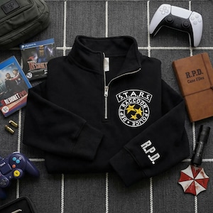 Könnte beinhalten: Schwarzes Sweatshirt mit Viertelreißverschluss, auf dem sich das S.T.A.R.S. Raccoon Police-Logo auf der Brust und R.P.D. auf dem Ärmel befinden. Das Bild zeigt Videospielartikel, darunter ein PlayStation 4-Spiel.