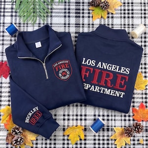 May include: A navy blue quarter-zip sweatshirt featuring the Los Angeles Fire Department logo on the left chest, the text Los Angeles Fire Department in red and white on the back, and the text Buckley 9-1-1 embroidered on the sleeve.