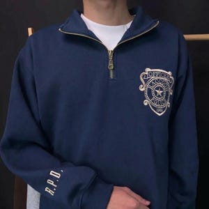 May include: A navy blue quarter-zip sweatshirt featuring a gold-tone zipper and a white embroidered Raccoon Police Officer badge on the chest. The right sleeve displays the embroidered text R.P.D. in a matching gold-tone thread color.