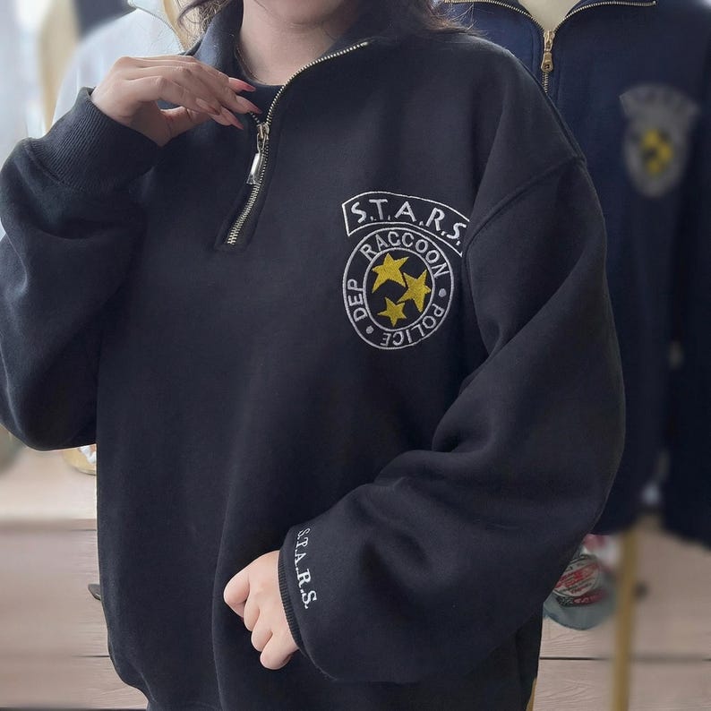 May include: A black quarter-zip sweatshirt featuring a white and yellow embroidered emblem on the chest that reads S.T.A.R.S. RACCOON POLICE DEP with three yellow stars. The left sleeve cuff includes the white embroidered text S.T.A.R.S.