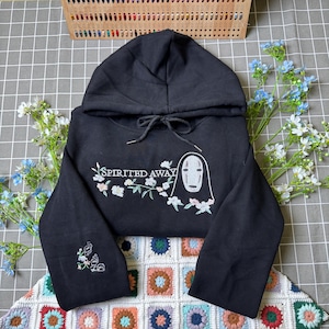 Pode incluir: Camisola preta com capuz com o texto "Spirited Away" e um design bordado de um personagem com detalhes florais. A camisola tem um capuz com cord&atilde;o e detalhes bordados na manga.