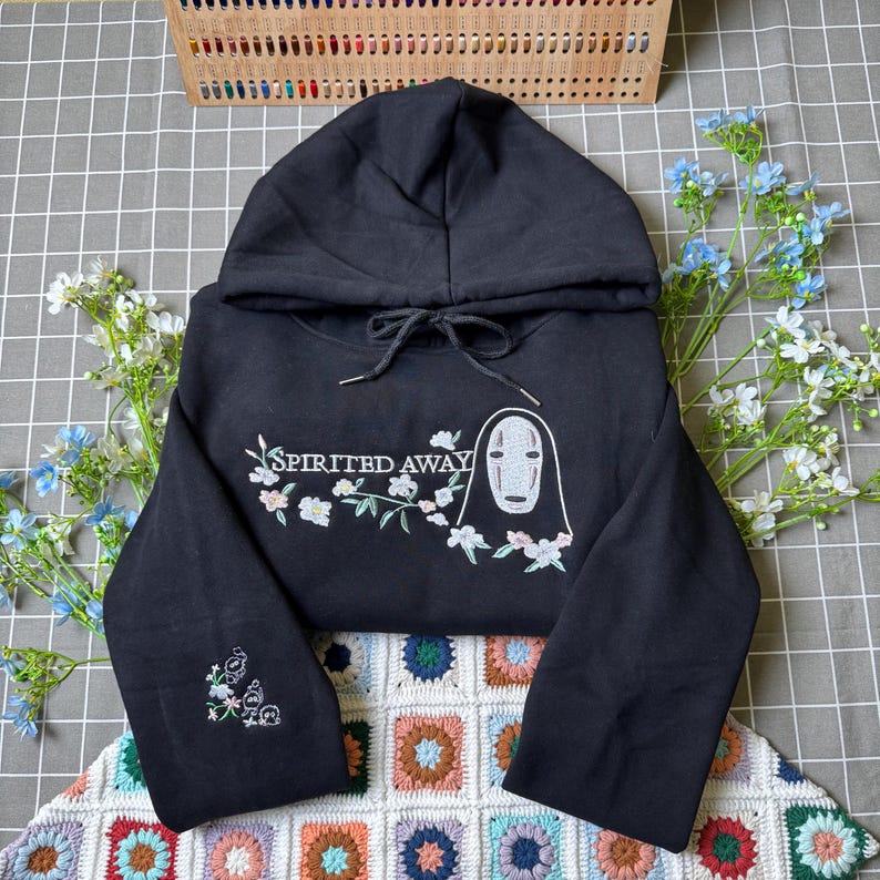 Pode incluir: Camisola preta com capuz com o texto "Spirited Away" e um design bordado de um personagem com detalhes florais. A camisola tem um capuz com cord&atilde;o e detalhes bordados na manga.