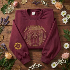 May include: A maroon sweatshirt featuring a gold geometric design and a smaller gold emblem on the left sleeve. The garment is folded on a wooden surface, decorated with flowers.