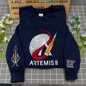 Op de afbeelding: Een marineblauw sweatshirt met ronde hals en een geborduurd Artemis II-missieontwerp op de borst, inclusief een maan, een raket en de tekst ARTEMIS II. De linkermouw heeft een raketafbeelding en de rechtermouw vermeldt de namen van de bemanning.
