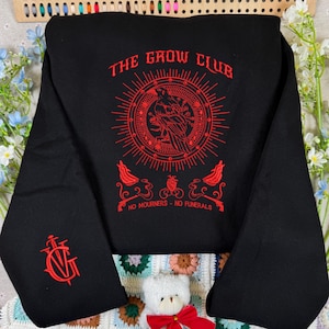 Op de afbeelding: Een zwart sweatshirt met lange mouwen en een rode grafische print. Het ontwerp bevat een centrale kraai in een cirkelvormige zonnestraal, de tekst THE CROW CLUB, een hart en de woorden NO MOURNERS NO FUNERALS. Een rood zwaardembleem staat op de mouw.