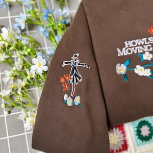 May include: Brown sweatshirt with embroidered design featuring a character and floral accents. The text "Howl's Moving Castle" is embroidered on the sleeve. The design includes red, white, and blue floral details.
