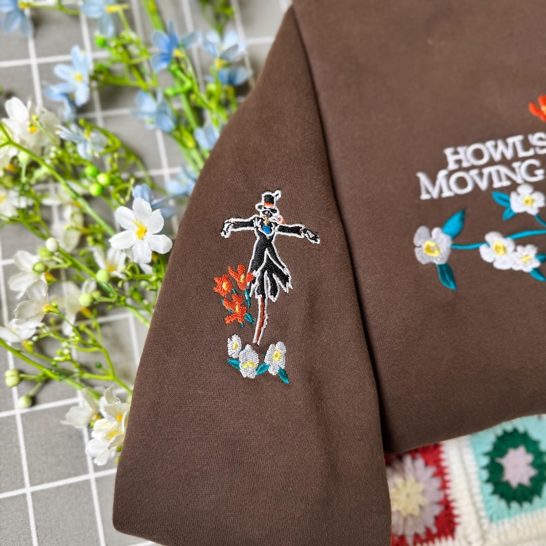 May include: Brown sweatshirt with embroidered design featuring a character and floral accents. The text "Howl's Moving Castle" is embroidered on the sleeve. The design includes red, white, and blue floral details.
