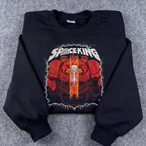 Peut inclure: Un sweat-shirt noir à col rond avec une broderie représentant une silhouette en armure rouge, centrée sur un élément de flammes verticales contenant un crâne et une colonne vertébrale blancs. Le texte SPACE KING est brodé en blanc au-dessus.