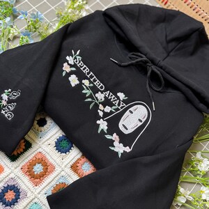 Pode incluir: Moletom preto com design floral e de personagem bordado. O design inclui o texto "SPIRITED AWAY" e um rosto de personagem. As mangas t&ecirc;m bordados florais. O moletom tem um capuz com cord&atilde;o.